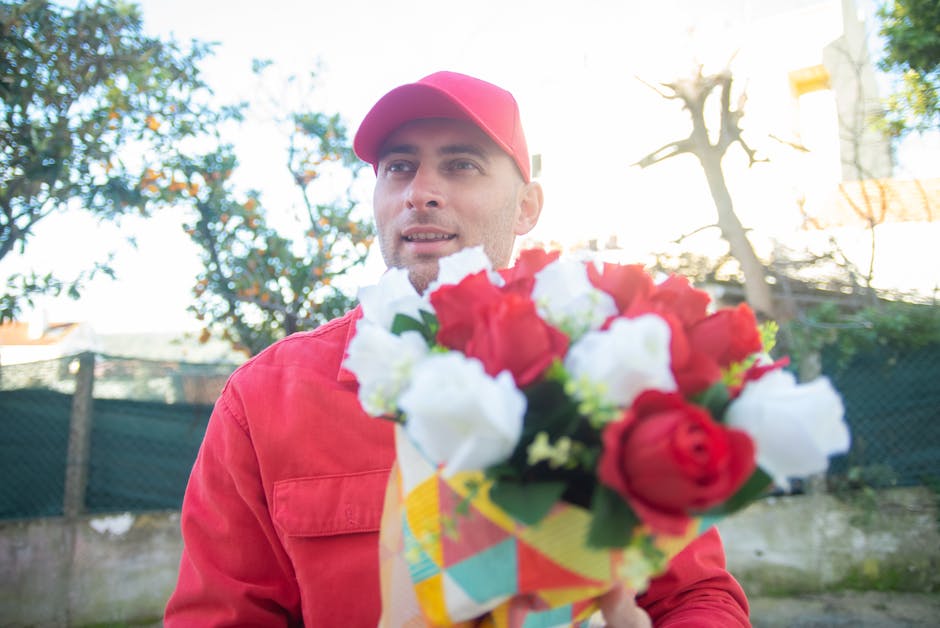 A man dressed in a red jacket and matching red cap holds a vibrant bouquet of fresh flowers, including red and white blooms, wrapped in colorful paper. He stands outdoors in a setting with trees, a fe