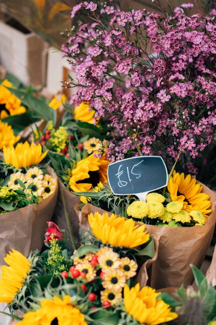 A vibrant display of fresh flowers arranged in brown paper wrapping, prominently featuring bright yellow sunflowers with large, round petals and dark centers, accompanied by smaller white and pink dai