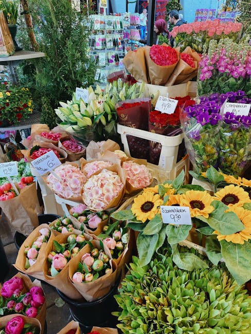 A vibrant display of fresh flowers at a florist stall, including white lilies with elongated petals and green stems, bright yellow sunflower heads with dark centers and broad green leaves, and purple 
