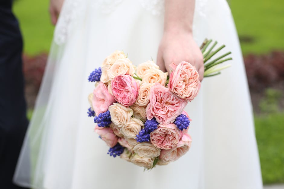 A bride holding a bouquet arrangement featuring pale pink and cream roses, complemented by clusters of vibrant blue berries, with fresh, velvety petals and sturdy green stems. The bouquet is wrapped i