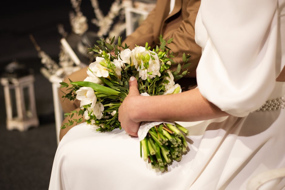 Close-up of a bride holding a bouquet of fresh white flowers, including lisianthus and freesias, accented with green foliage. The bouquet is wrapped with a white lace ribbon and rests on her lap, drap