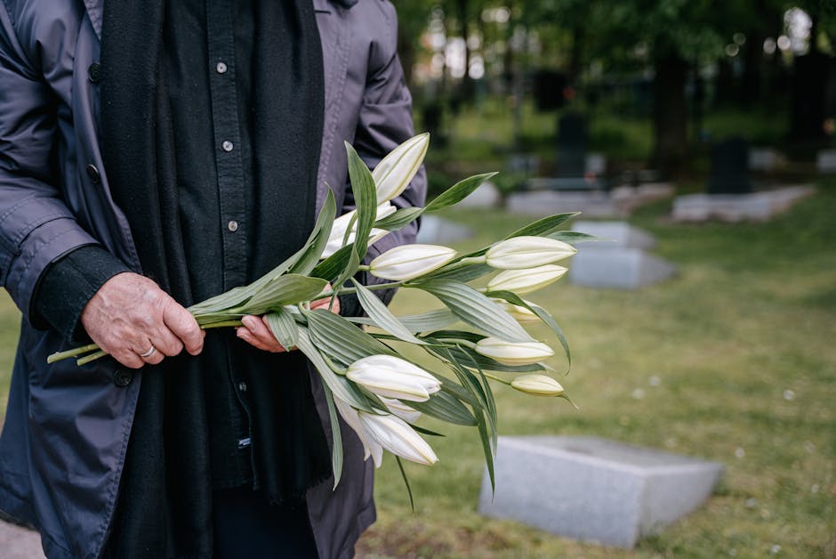 A person dressed in dark clothing and a coat is holding a bouquet of large white lilies with long, green leaves. The bouquet is unwrapped, and the lilies' petals are smooth and slightly curved, indica