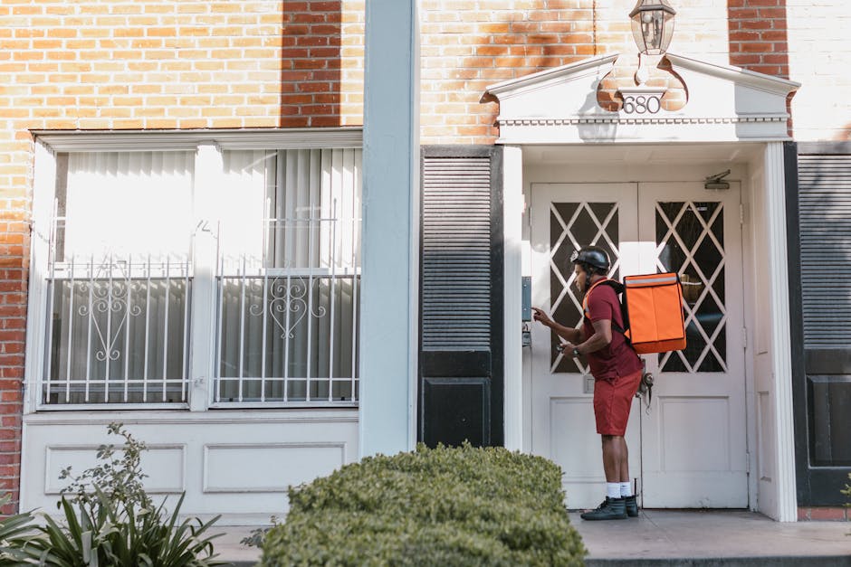 A delivery person in a red uniform and helmet is interacting with the front door of a residential building, holding a smartphone in one hand and using the other to unlock or open the door, which has a