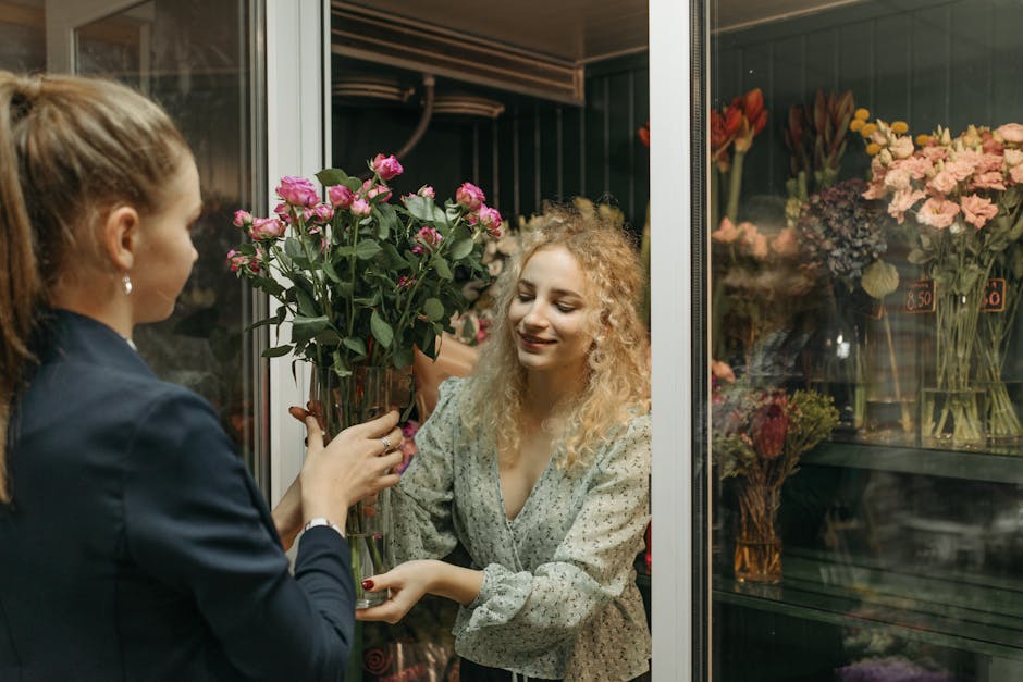 A florist at flowerdeliverykennington.co.uk is handing a tall, clear glass vase filled with a bouquet of vibrant pink roses and green foliage to a smiling customer inside a floral shop. The bouquet fe