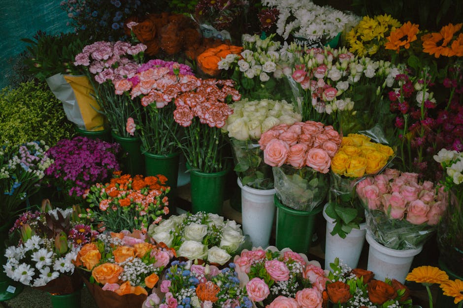 A vibrant display of fresh flower bouquets and potted plants arranged in green and white containers inside a floral shop. The arrangement includes pink, orange, white, yellow, and purple flowers, such